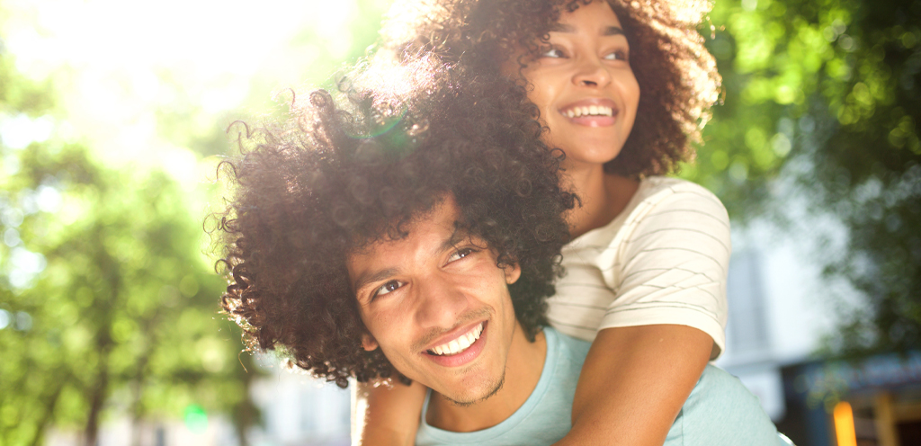 young african american couple smiling outside with man holding woman in piggyback position looking to the right